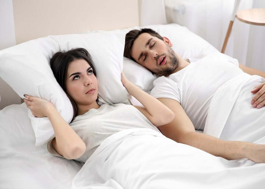 Couple in bed, man snoring loudly, woman covering ears, illustrating need for anti-snoring solutions like Nitetronic smart pillow.