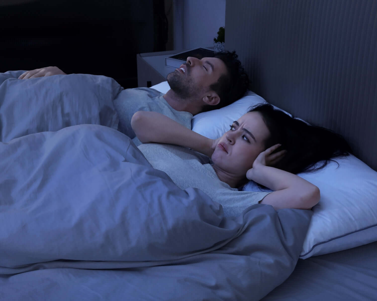 Couple in bed, man snoring while woman struggles to sleep, highlighting the impact of snoring on sleep quality and well-being.