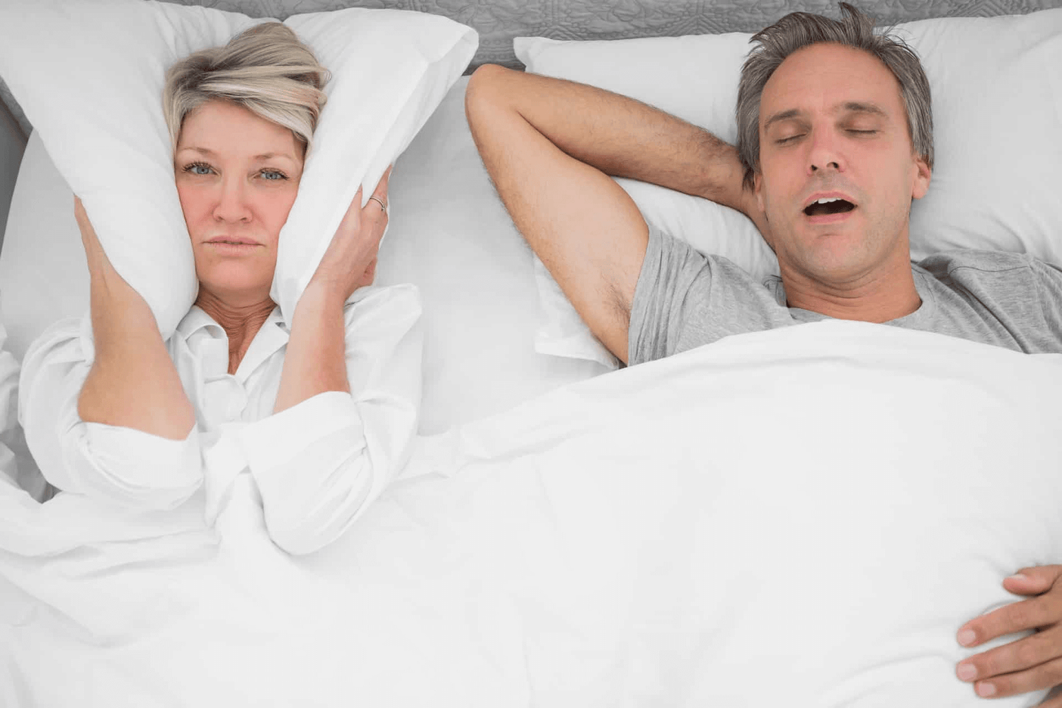 Man snoring in bed next to frustrated woman covering ears with pillow, highlighting sleep apnea issues.