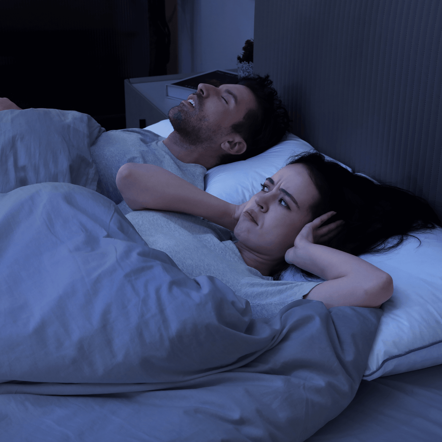 Couple in bed with woman covering ears, disturbed by snoring partner, illustrating snoring-related sleep issues.