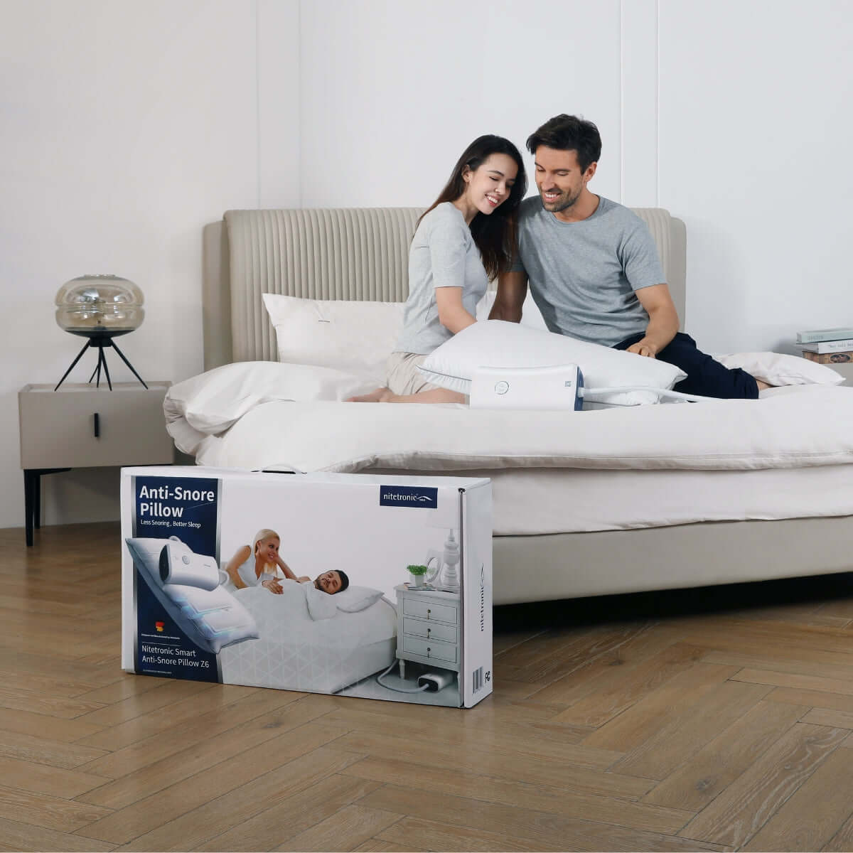 Couple using Nitetronic Anti-Snore Pillow on bed, enhancing sleep quality by addressing snoring issues, product box in foreground.