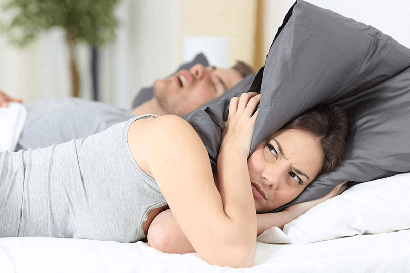 Woman covers ears with pillow while man snores, highlighting sleep disturbance issues.