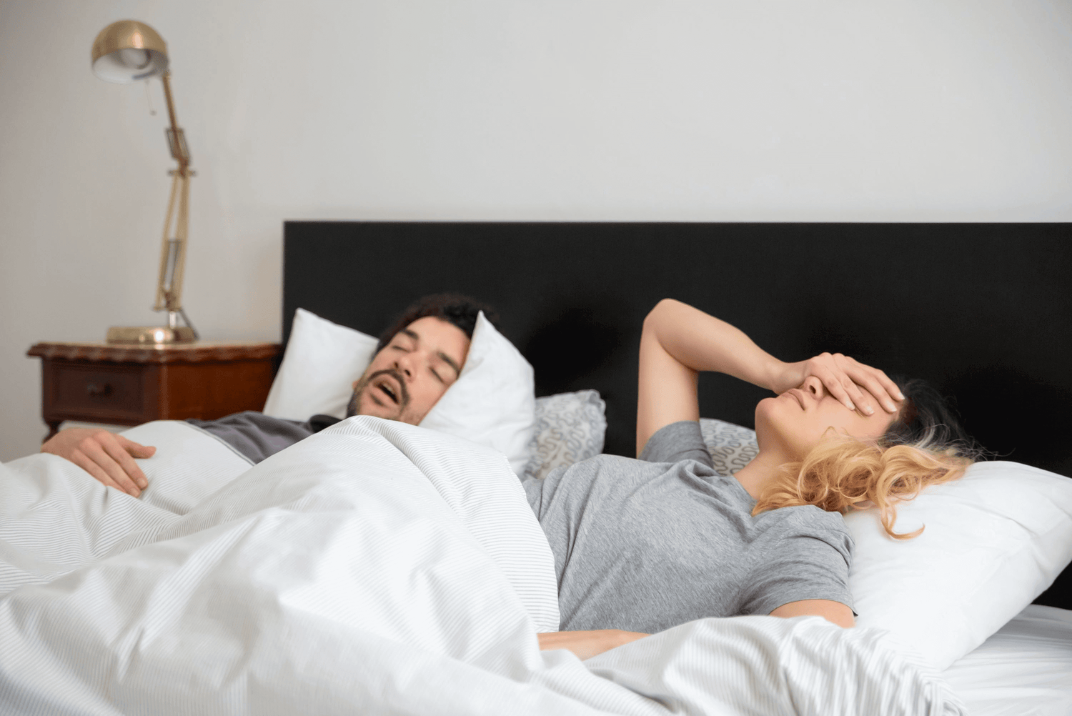 Couple in bed, man snoring, woman looking frustrated; illustrating sleep apnea issues requiring the right pillow for better sleep.