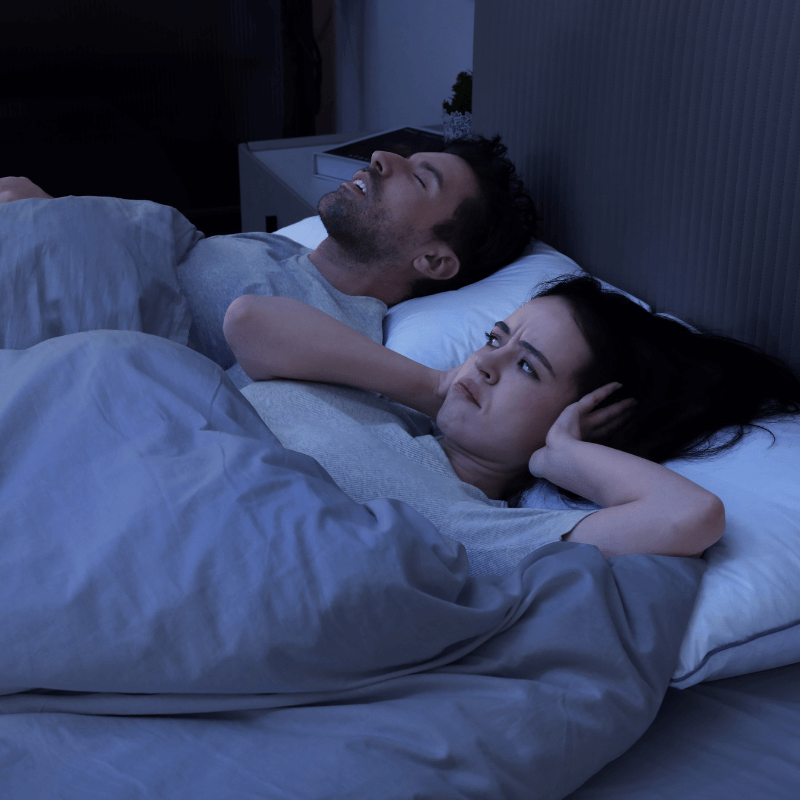Man snoring loudly in bed while woman covers her ears, illustrating sleep disturbance due to snoring.