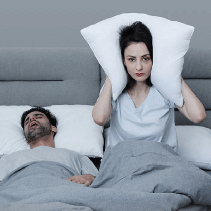 Couple in bed, woman covering ears with pillow to block snoring partner. Explore snoring causes and remedies for better sleep.