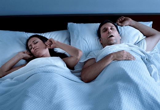 Couple sleeping in bed, man snoring, woman covering ears, illustrating snoring impact on sleep.