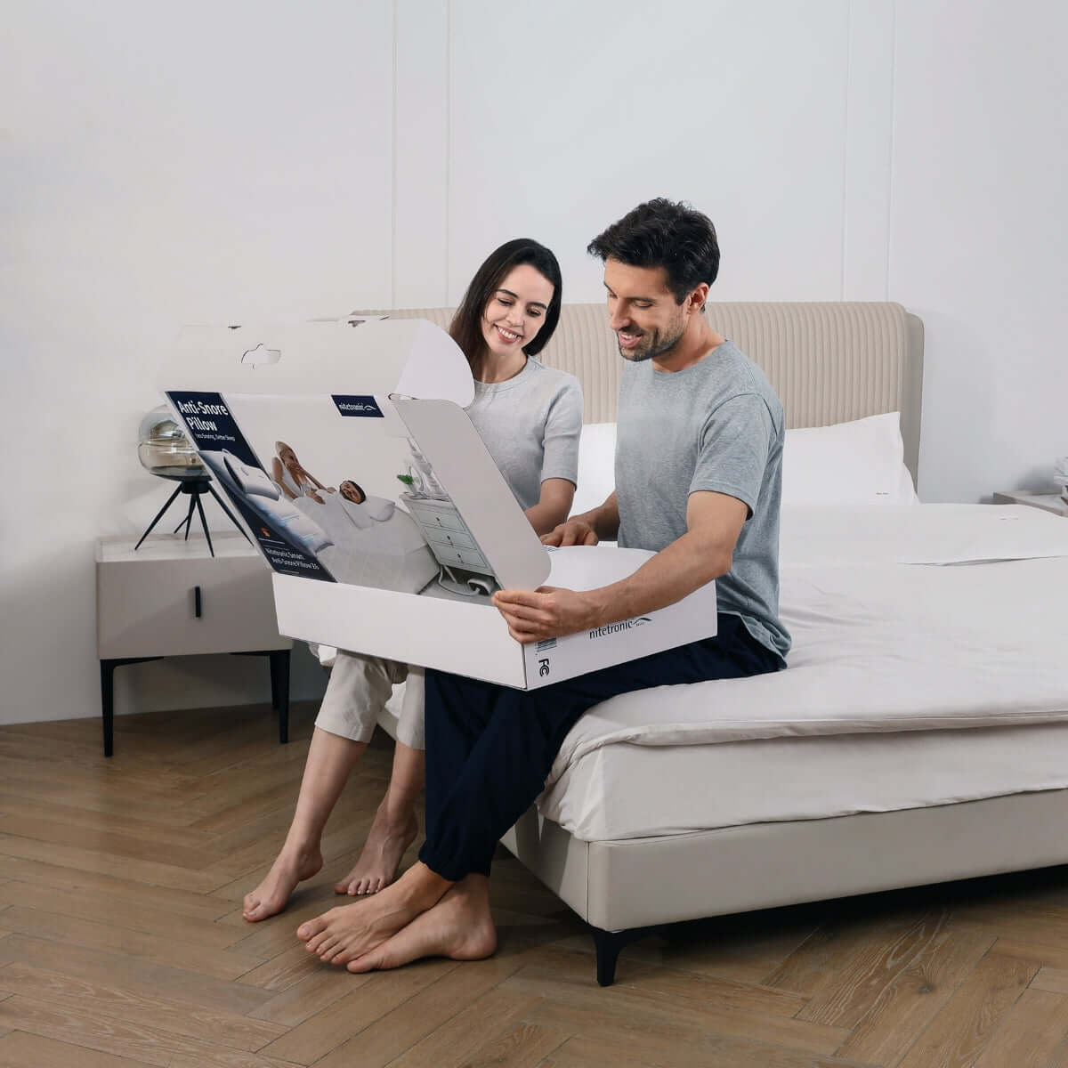 Couple unboxing an anti-snoring pillow on a bedroom bed, looking excited