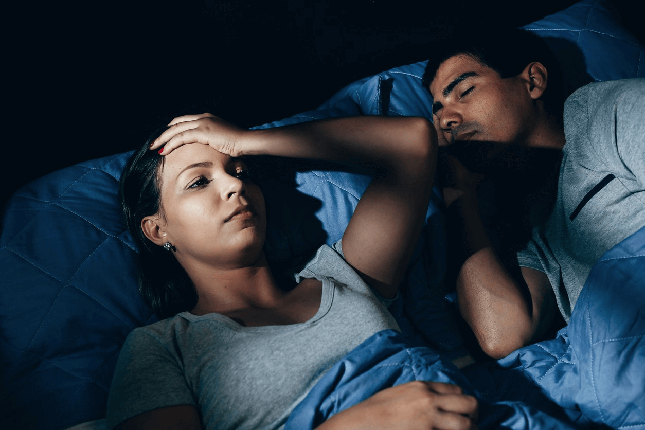 Woman lying awake next to sleeping partner, highlighting the challenge of snoring in sleep.