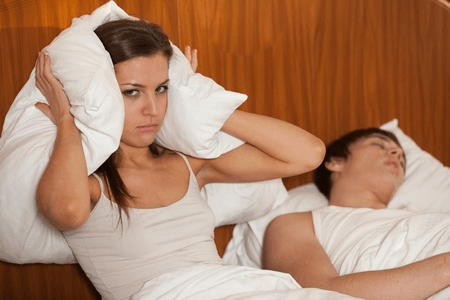 Woman covering her ears with a pillow next to a snoring partner, highlighting the need for an anti-snoring pillow solution.