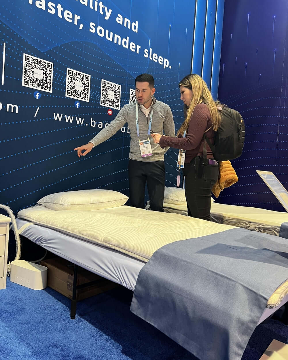 Two people discussing features at a mattress exhibit, showcasing comfortable sleep solutions with a model bed on display.