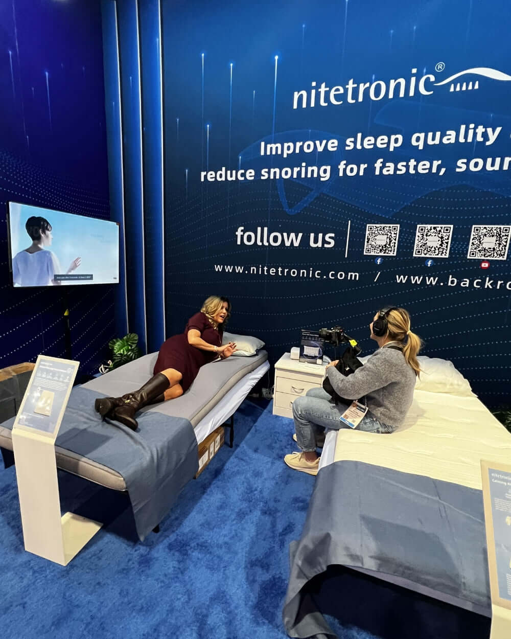 A person demonstrating a sleep product on a blue bed at a Nitetronic stand at an exhibition, with another person filming the demonstration.