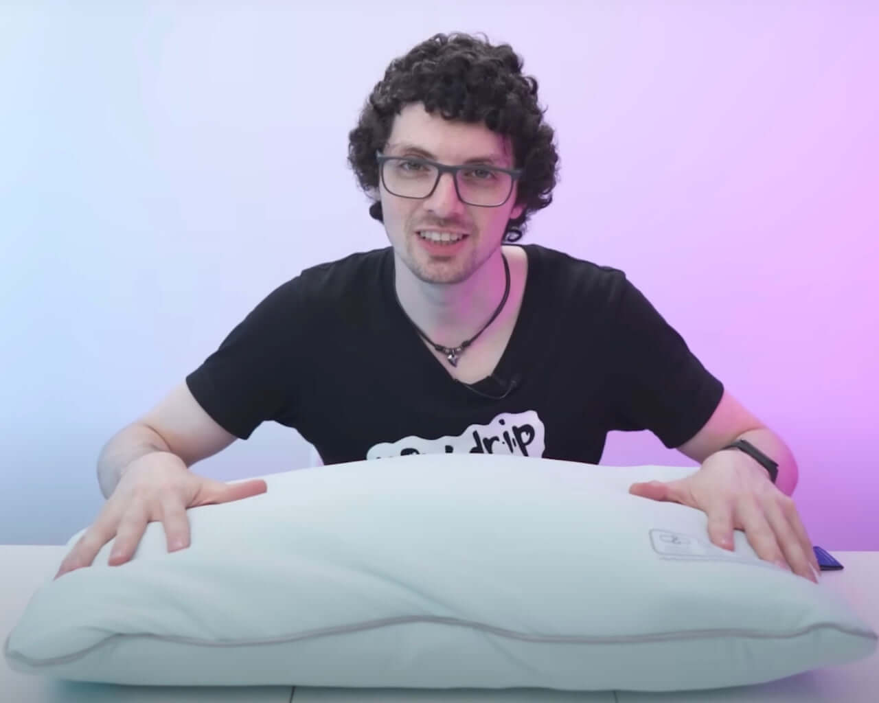 Person demonstrating a comfortable white pillow on a table under cool lighting, wearing glasses and a black t-shirt with print.