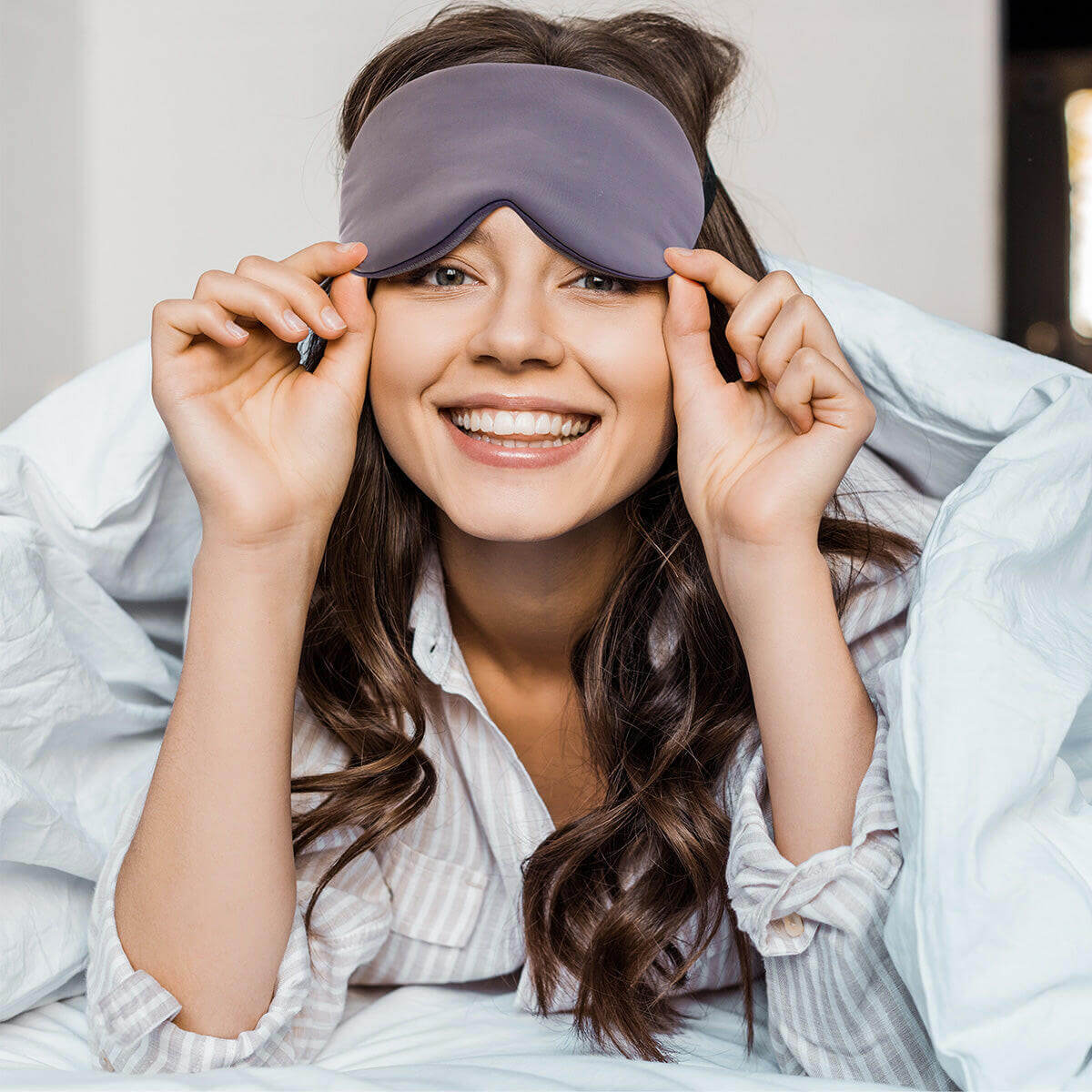 Woman smiling while wearing Nitetronic Sleep Mask in bed, demonstrating ergonomic design and light-blocking features for better rest.