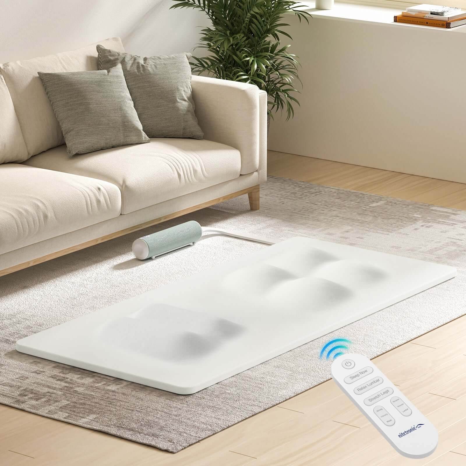 White massage mat with remote control on a light rug beside a beige sofa in a modern living room setting.